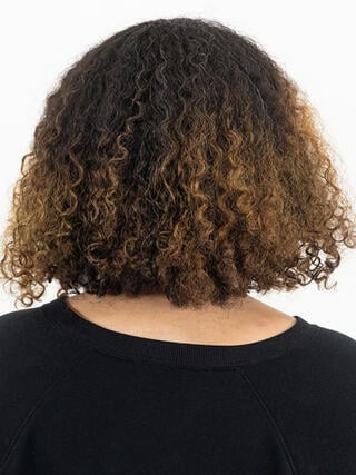 Foto "antes": vista trasera de una mujer con cabello negro corto y rizado con las raíces crecidas antes de la coloración.